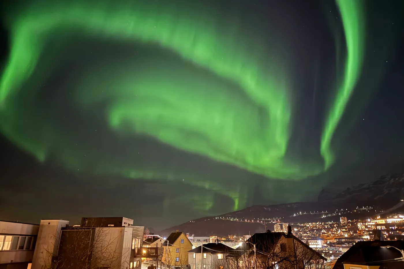 Brillante aurora boreale verde che si inarca sulle case illuminate e sulle luci del pendio di Narvik di notte