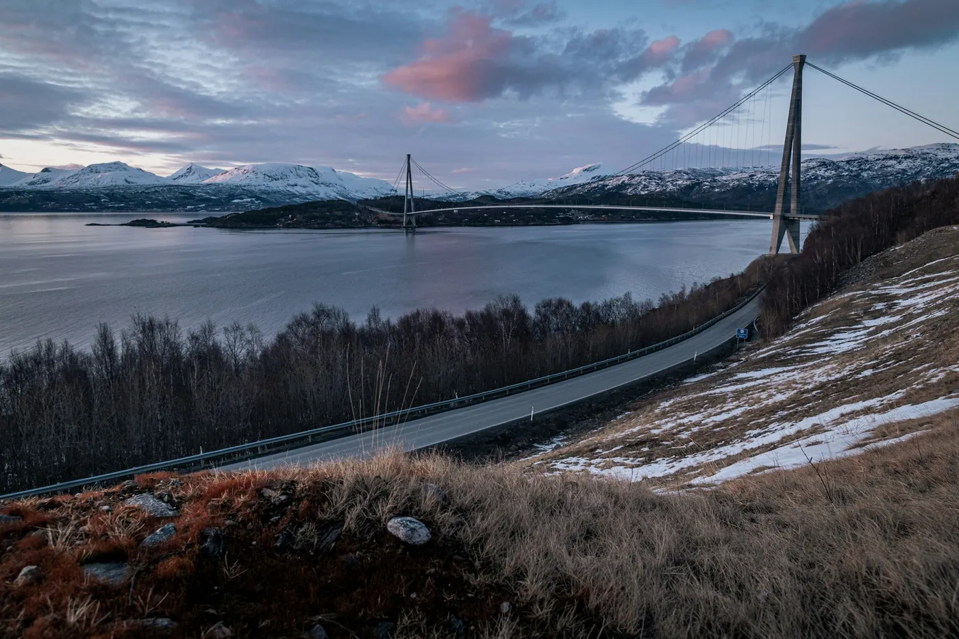 Long pont suspendu qui franchit un fjord, route d’accès qui serpente sur un versant d’herbe sèche et de neige par plaques, nuages roses