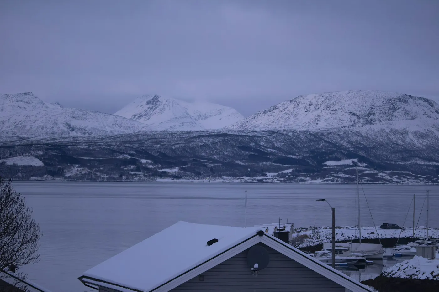 Toits enneigés et petit port de plaisance au premier plan, large fjord et chaîne de montagnes enneigées derrière, sous un ciel d’hiver bleu-gris