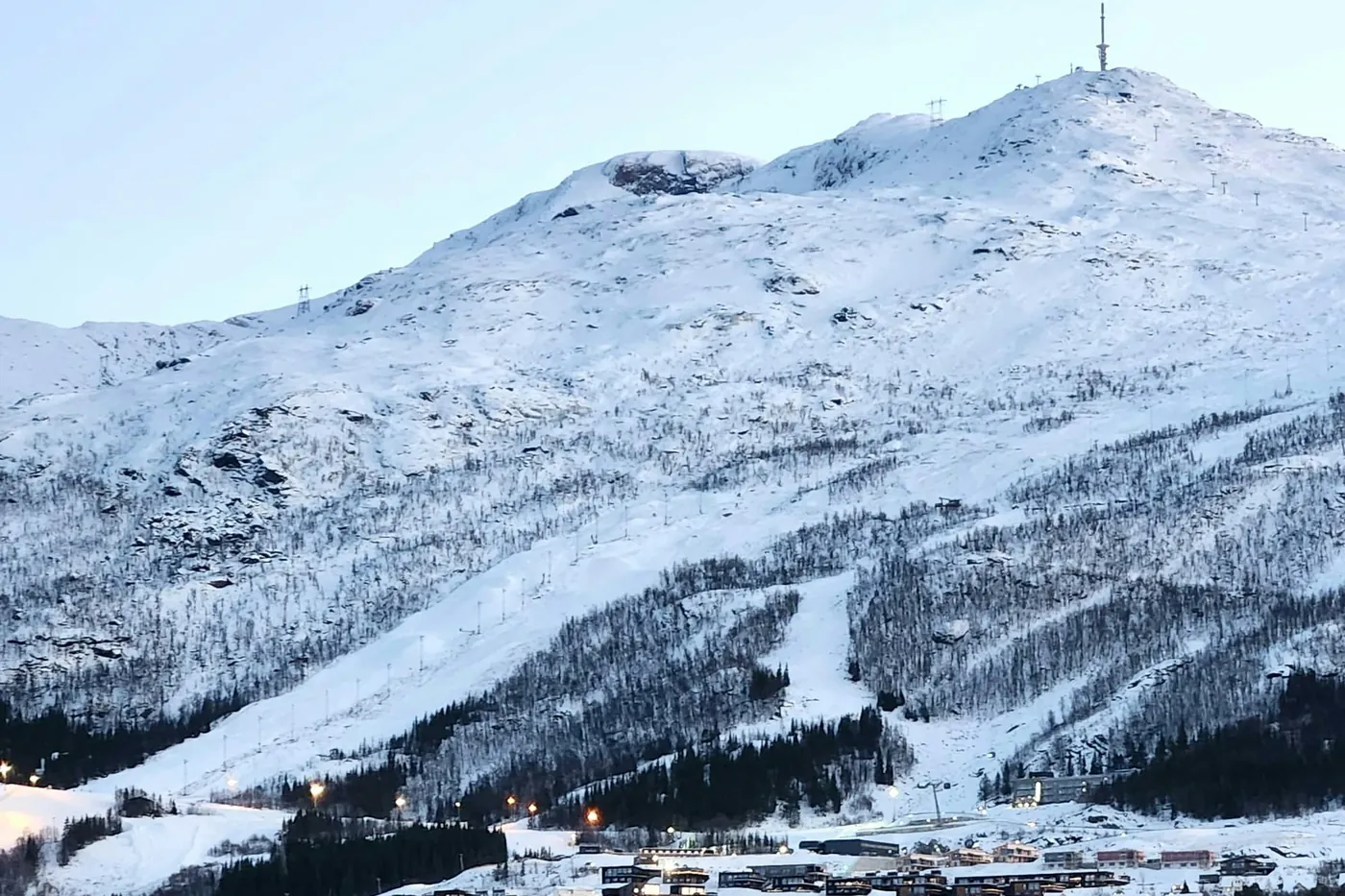 Narvikfjellet nevado con pistas de esquí, torres de remonte y un mástil en la cima, y edificios de la ciudad a sus pies