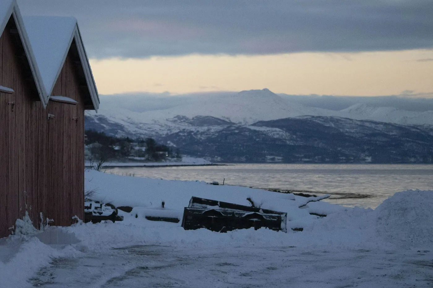 Fila di rimesse rosse in legno con i tetti innevati sulla riva a Narvik, dietro il fiordo e montagne innevate sotto un pallido cielo serale