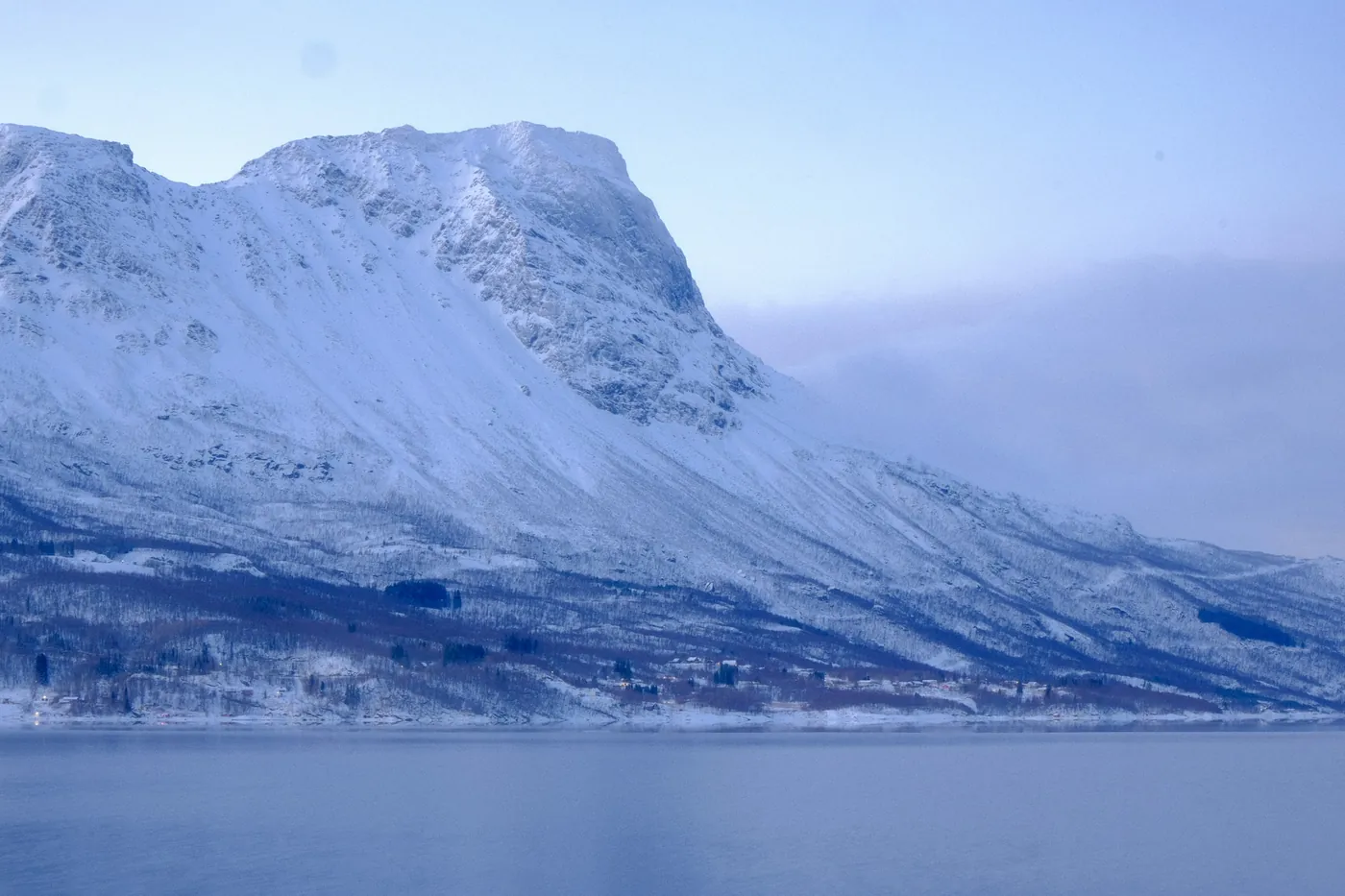 Un’imponente montagna coperta di neve che si alza ripida dall’acqua calma del fiordo vicino a Narvik sotto un pallido cielo azzurro