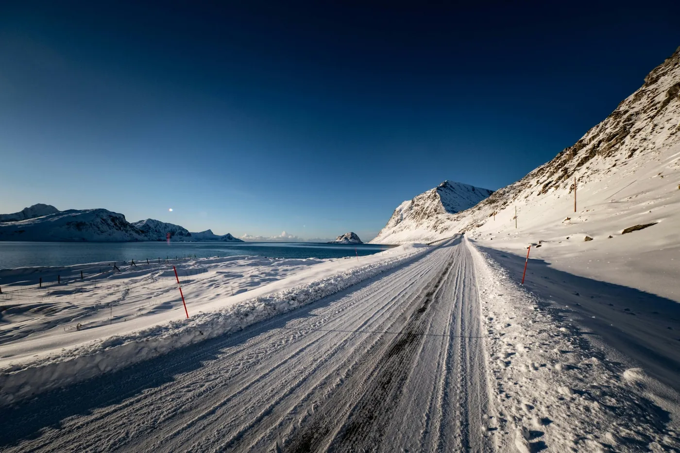 Carretera costera vacía con nieve compactada y postes rojos de señalización junto al mar, con colinas nevadas bajo un cielo azul intenso