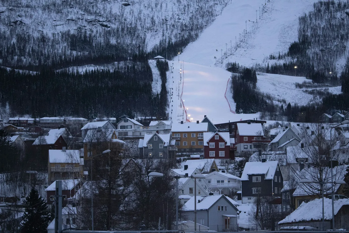 Una pista de esquí iluminada con focos que atraviesa un bosque de abedules sobre casas cubiertas de nieve en Narvik