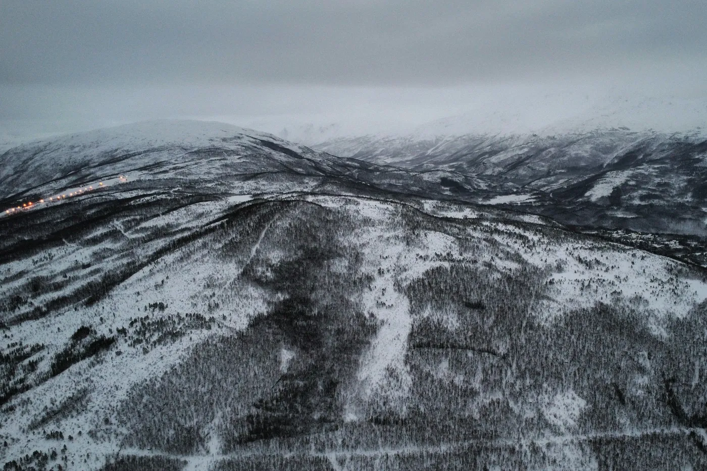 Veduta aerea di pendii montuosi innevati e boscosi in una giornata grigia, un treno illuminato visibile come una linea di luci sul versante lontano