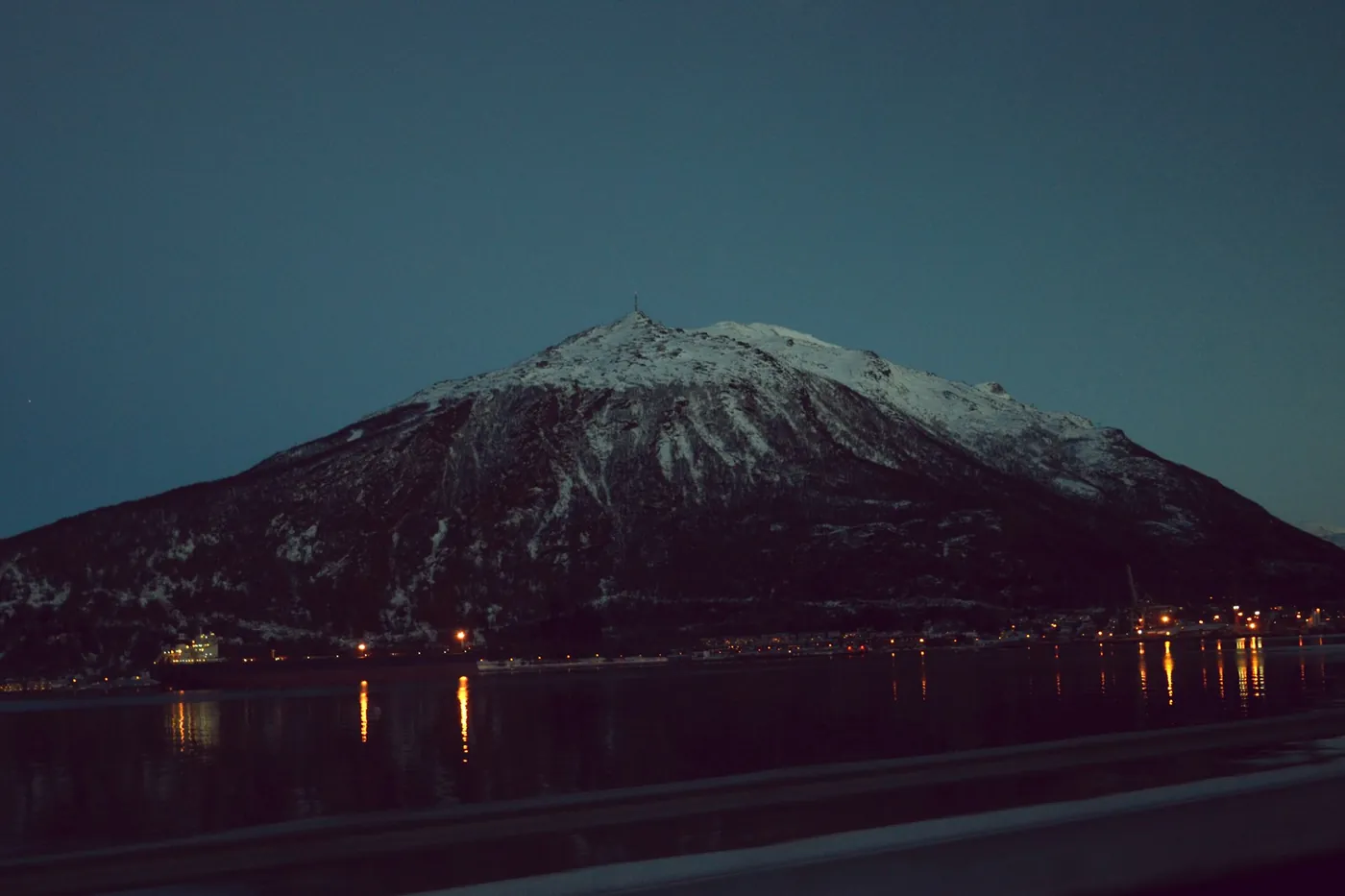 Montagna scura con una vetta innevata al crepuscolo, piccole luci della città riflesse nel fiordo